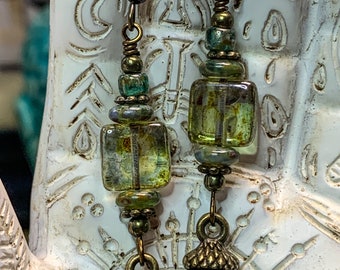 May include: A pair of dangle earrings featuring bronze-toned metal acorn charms at the base. Each earring includes a square, translucent green glass bead accented with smaller metallic and iridescent beads on a bronze-toned wire hook.
