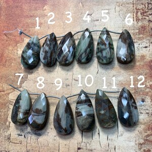 May include: Twelve teardrop-shaped gemstone beads with a brown, white, and green marbled pattern. The beads are strung on a silver wire and numbered 1 through 12.