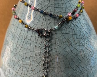 May include: A delicate necklace featuring a thin dark metal chain interspersed with small, multi-colored faceted gemstone beads in shades of pink, yellow, green, and blue. The necklace includes a lobster claw clasp and an adjustable chain extender.
