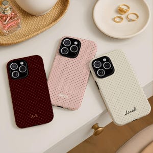 May include: Three phone cases with a polka dot pattern. One is burgundy with "A+E" at the bottom, one is pink with "oliva" at the bottom, and one is cream with "Sarah" at the bottom. Gold rings and a perfume bottle are also visible.