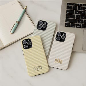 May include: Three phone cases with vertical striped patterns in yellow, light green, and off-white. The yellow case has the initials "sgb", and the off-white case has "aMe". A pen and notebook are in the background.