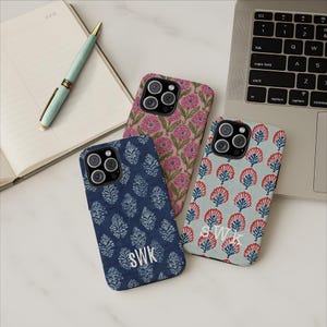 May include: Three phone cases with floral and leaf patterns in pink, blue, and light blue. One case has the initials "SWK" in white. A pen and notebook are in the background, along with a laptop.