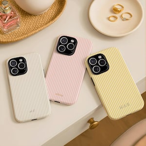 May include: Three striped phone cases in cream, pink, and yellow. The cream case has the initials "A.E." and the pink case has the name "olivia" printed on it. The yellow case has the initials "MAE". Gold rings and a perfume bottle are also visible.