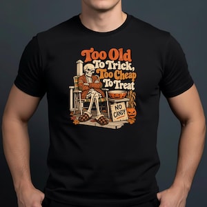 May include: A black t-shirt featuring a graphic of a skeleton sitting in a chair wearing a cardigan and slippers. The text reads, "Too Old To Trick, Too Cheap To Treat" and "NO CANDY" on a sign next to a pumpkin and a bowl of candy.