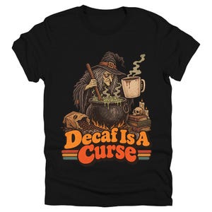 May include: A black t-shirt featuring a graphic illustration of a witch stirring a bubbling cauldron with a wooden spoon. Beside the cauldron are coffee beans, a mug of steaming coffee, books, and a skull. The text reads Decaf Is A Curse.