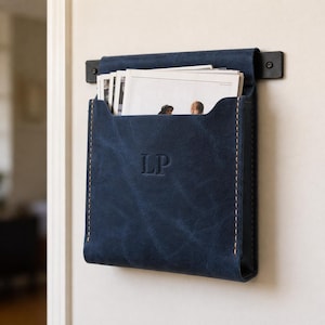 May include: A navy blue leather wall-mounted organiser with the initials "LP" embossed on the front. The organiser is filled with papers and has contrasting beige stitching. It is attached to the wall with a black metal bracket.