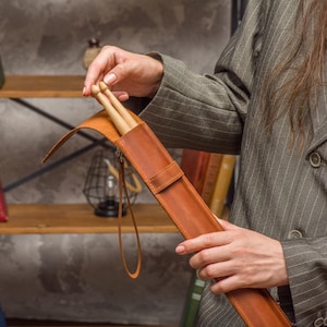 Puede incluir: Un estuche de baquetas de cuero marrón con una correa. El estuche está abierto y hay dos baquetas de madera en su interior.