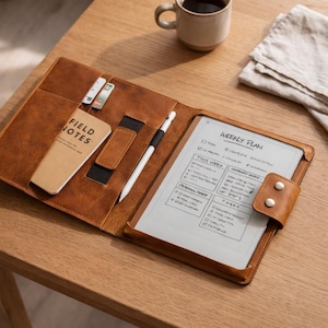 Puede incluir: Un folio de cuero marrón con una tableta, un bloc de notas y un bolígrafo sobre una mesa de madera. La tableta muestra un plan semanal. Un pequeño cuaderno Field Notes y tarjetas están en los bolsillos del folio. Una taza de café está cerca.