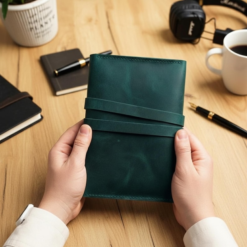 Op de afbeelding: Een donkergroen leren notitieboek met een wikkelriemsluiting. Het notitieboek wordt in handen gehouden, met andere notitieboekjes, een pen en een koffiekopje op de achtergrond. Het notitieboek heeft een rijke, effen kleur.