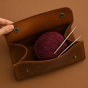 May include: A brown leather knitting project bag with a burgundy ball of yarn and two silver knitting needles inside.