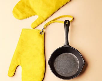 May include: Overhead shot of a black cast iron skillet with a long handle, next to two bright yellow oven mitts. The mitts are made of a soft material and have a loop for hanging. The skillet is small, perfect for individual servings.