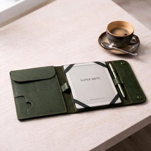 May include: A dark green leather folio case, open to reveal a tablet with "SUPER NOTE" printed on it. A silver pen is held in a loop. A cup of coffee on a saucer is in the background. The case rests on a light wooden surface.
