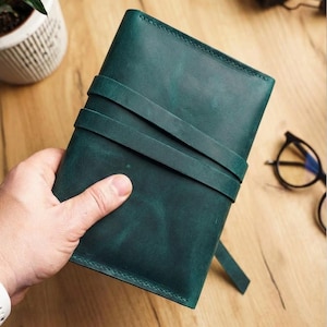 Op de afbeelding: Een turquoise leren notitieboek met een wikkelriemsluiting. Het notitieboek wordt in een hand gehouden, waarbij de textuur en kleur van het leer worden getoond. Het ontwerp is eenvoudig en elegant, geschikt om in te schrijven of te schetsen.