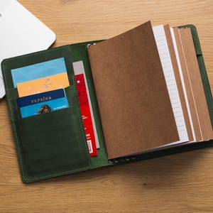 May include: A green leather bound notebook with a brown paper cover and lined pages. The notebook has several pockets on the front cover, containing a passport, cards, and other documents.