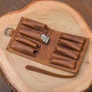 May include: A brown leather roll-up case featuring multiple stitched slots for storage, a silver metal buckle closure on the left side, and a long leather strap. One slot contains a small silver harmonica with visible metal components.