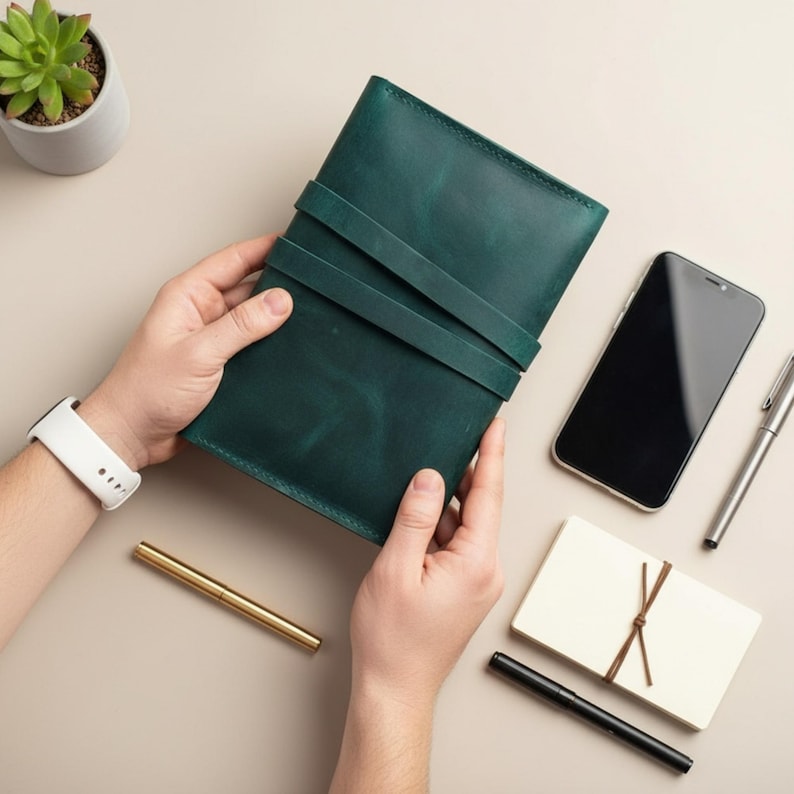Op de afbeelding: Een donkergroen leren notitieboek met twee bandjes, vastgehouden door handen. Een witte smartwatch, een gouden pen, een zwarte smartphone, een zilveren pen, een klein notitieblok en een vetplant in een pot zijn ook zichtbaar.