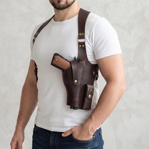 Könnte beinhalten: Ein dunkelbraunes Schulterholster aus Leder mit sicherem Gurtsystem, messingfarbenen Metallbeschlägen und einer integrierten Tasche, auf deren Vorderklappe die Initialen LK eingeprägt sind. Das Holster enthält eine Handfeuerwaffe mit Holzgriff.