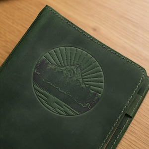 May include: A dark green leather journal with a circular embossed design of a mountain range and sun rays.