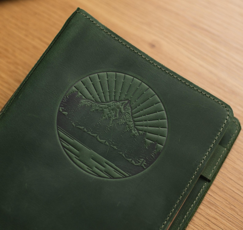May include: A dark green leather journal with a circular embossed design of a mountain range and sun rays.