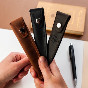 May include: Three leather pen cases in brown, black, and dark brown. The cases are made of a textured leather and have a snap closure. The dark brown case has the initials "SGA" embossed on the front.