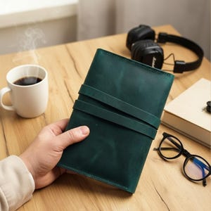 Op de afbeelding: Een donkergroen leren notitieboek met twee bandjes, vastgehouden door een hand. Een witte koffiemok met stoom staat links. Zwarte koptelefoon, een boek en een bril liggen op het houten oppervlak.