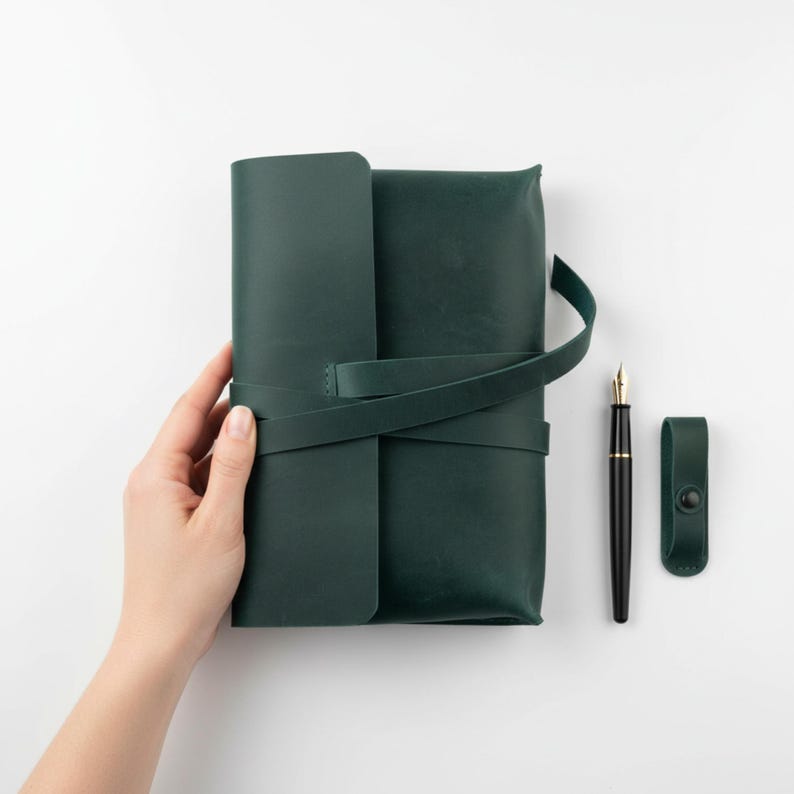 Op de afbeelding: Een donkergroen leren notitieboek met een wikkelriem, een zwarte vulpen met een gouden penpunt en een bijpassende penhouder. Het notitieboek is gesloten en heeft een minimalistisch ontwerp.