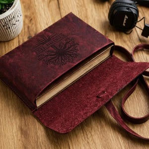Puede incluir: Un diario de cuero burdeos con un diseño de árbol y las palabras "CUSTOM TEXT" grabadas en la portada. El diario está abierto, revelando las páginas en el interior. Se adjunta una correa de cuero a juego. Hay unos auriculares en el fondo.
