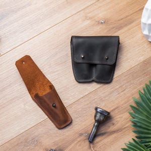 May include: Two leather cases for musical instruments. One is brown and the other is black. A silver trumpet mouthpiece is also in the image.