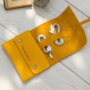 May include: Mustard yellow leather cable organizer with a flap closure and two silver snaps. The organizer holds two white charging cables. The text "DonskyStore" is printed on the flap.