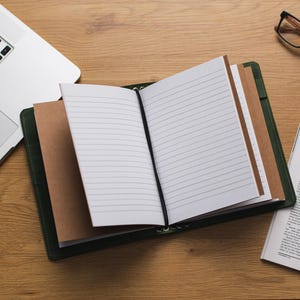 May include: A green leather bound journal with blank pages and a brown paper bookmark. The journal is open and lying on a wooden table with a laptop and a pair of glasses.