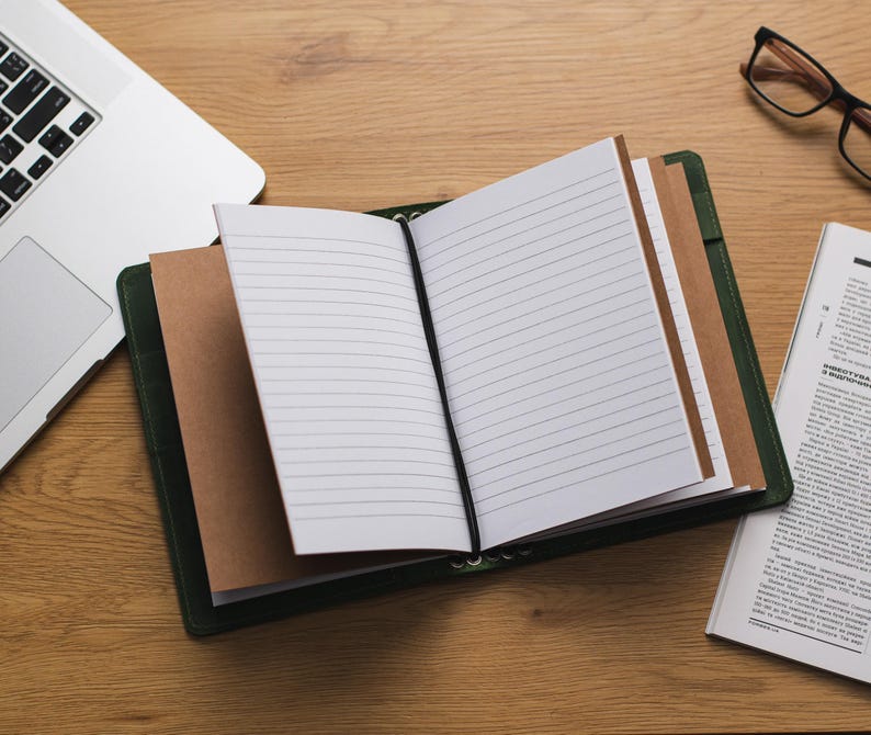 May include: A green leather bound journal with blank pages and a brown paper bookmark. The journal is open and lying on a wooden table with a laptop and a pair of glasses.