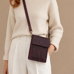 Puede incluir: Un bolso bandolera de cuero color morado oscuro con cierre de solapa rectangular y correa ajustable a juego. El bolso cuenta con herrajes metálicos en tono latón antiguo, incluyendo clips giratorios y remaches, y tiene un acabado mate.