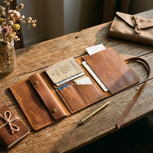 May include: Open leather passport holder with multiple pockets, holding a passport, cards, and papers. The holder is open on a wooden surface, with a pen and a small notebook tied with twine nearby. A matching leather envelope is in the background.