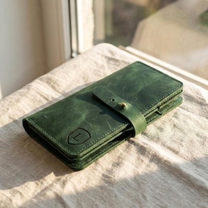 Puede incluir: Una cartera rectangular de cuero verde bosque con acabado mate liso y cierre de correa asegurado con un pequeño perno de metal color latón. La parte frontal de la cartera muestra un emblema de escudo estampado con la letra L.