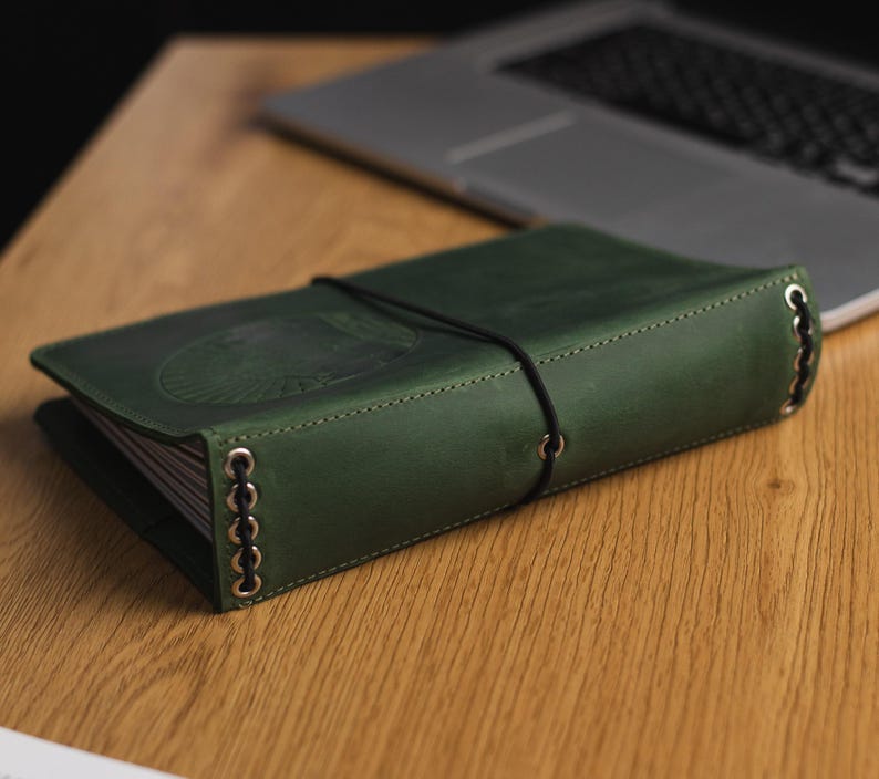 May include: A dark green leather-bound journal with a black elastic band closure and silver metal accents. The journal is closed and lying on a wooden surface.