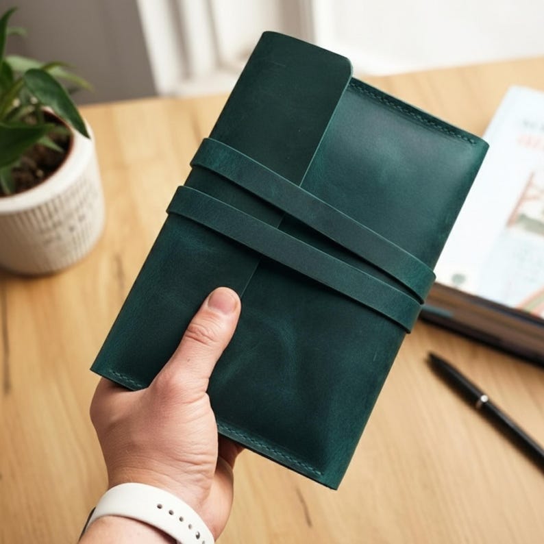 Op de afbeelding: Een donkergroen leren notitieboek met een wikkelriemsluiting. Het notitieboek is rechthoekig en lijkt handgemaakt. De omslag heeft een effen, diepgroene kleur. Het notitieboek wordt in de hand van een persoon gehouden.