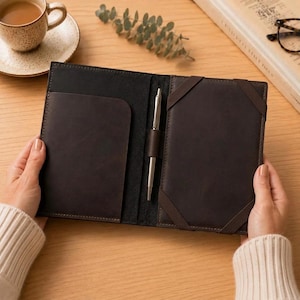 Puede incluir: Una funda de cuero marrón oscuro para cuaderno con un portalápices vertical en el centro, un bolsillo en el lado izquierdo y un panel derecho con dos bandas elásticas en las esquinas para asegurar un cuaderno. El interior está forrado en negro.