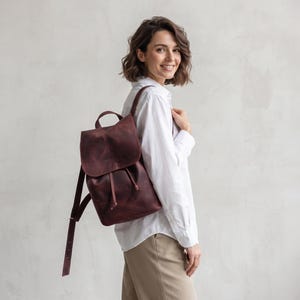 May include: A dark burgundy leather backpack featuring a fold-over flap with a drawstring closure, a top handle, and adjustable shoulder straps. The bag has a smooth, matte finish and is worn over one shoulder, showcasing its vertical structure.