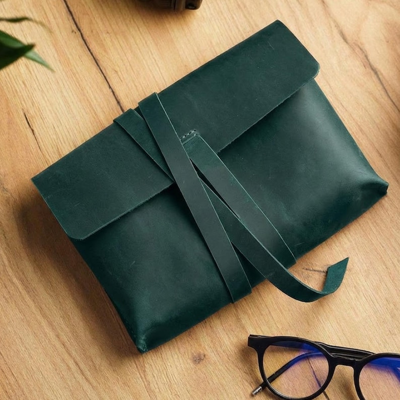 Op de afbeelding: Een donkergroene leren clutch met een klep en een riemsluiting. De rechthoekige clutch lijkt handgemaakt. Een bril met zwart montuur ligt op het houten oppervlak naast de clutch.