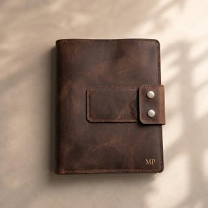 May include: A dark brown leather notebook cover featuring a rectangular front pocket and a side strap secured with two silver-toned metal snaps. The bottom right corner of the cover is embossed with the capital letters MP in a serif font.