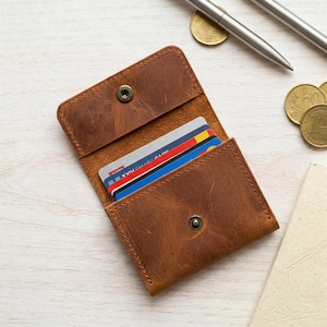 May include: A brown leather card holder featuring a snap button closure and a flap design. The wallet contains several credit cards with visible blue, red, and white accents. The leather has a textured, distressed finish with visible grain.