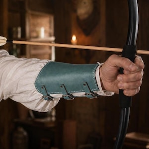 May include: A teal leather forearm guard featuring three adjustable metal buckles and white stitching, worn over a cream-colored long-sleeved shirt. The guard is positioned on the arm while holding a black bow with a wooden arrow nocked.