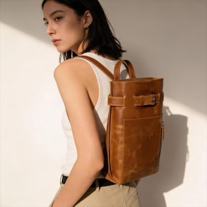 May include: A brown leather backpack with a vertical design, featuring a top handle, adjustable straps, and a front pocket with a zip. The bag has a buckle closure and is worn over the shoulders.