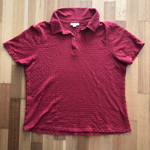 Könnte beinhalten: Ein kurzärmeliges, rotes Poloshirt mit strukturiertem Muster. Das Shirt hat einen Kragen und eine Knopfleiste mit drei Knöpfen. Der Stoff scheint ein strukturiertes Strick zu sein. Das Shirt wird auf einer Holzoberfläche präsentiert.