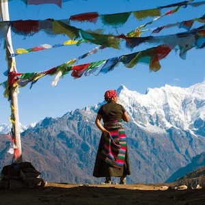 May include: A woman in a colorful traditional dress stands on a mountaintop, looking out at a snow-capped mountain range. Prayer flags hang in the wind, creating a vibrant scene.