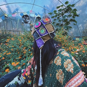 May include: A patchwork beanie with earflaps featuring brown, purple, and grey fabric panels adorned with circular embroidered accents in orange, pink, and green. The hat includes a top pom-pom and braided tassels hanging from the earflaps.