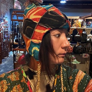 May include: A multi-colored patchwork hat featuring a top pom-pom and two long braided side tassels. The hat is constructed from various fabric segments in shades of red, black, orange, and teal, joined by visible stitching. The hat covers the ears.