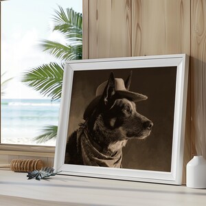 May include: A sepia-toned framed print of a dog wearing a cowboy hat and scarf. The dog is in profile, facing right, with a serious expression. The white frame is set on a white surface near a window with a beach view.