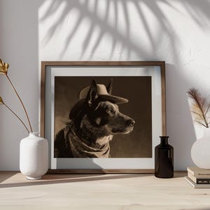May include: Sepia-toned framed print of a dog wearing a cowboy hat and scarf. The dog is a medium-sized breed with dark fur, set against a neutral background. The print is displayed on a wooden surface with decorative vases and books.