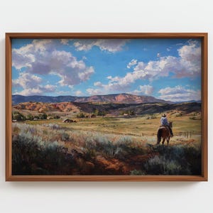 May include: A framed landscape painting depicts a cowboy on horseback in a vast, grassy field. The artwork features a bright blue sky with fluffy white clouds, rolling hills, and a rustic wooden frame.